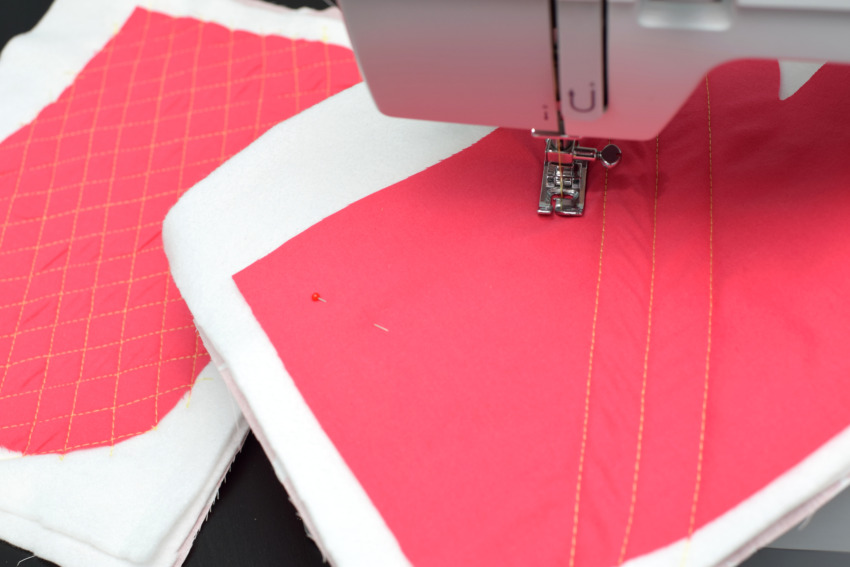 A close-up of a sewing machine needle stitching yellow diagonal diamond patterns onto pink fabric and thick batting layers